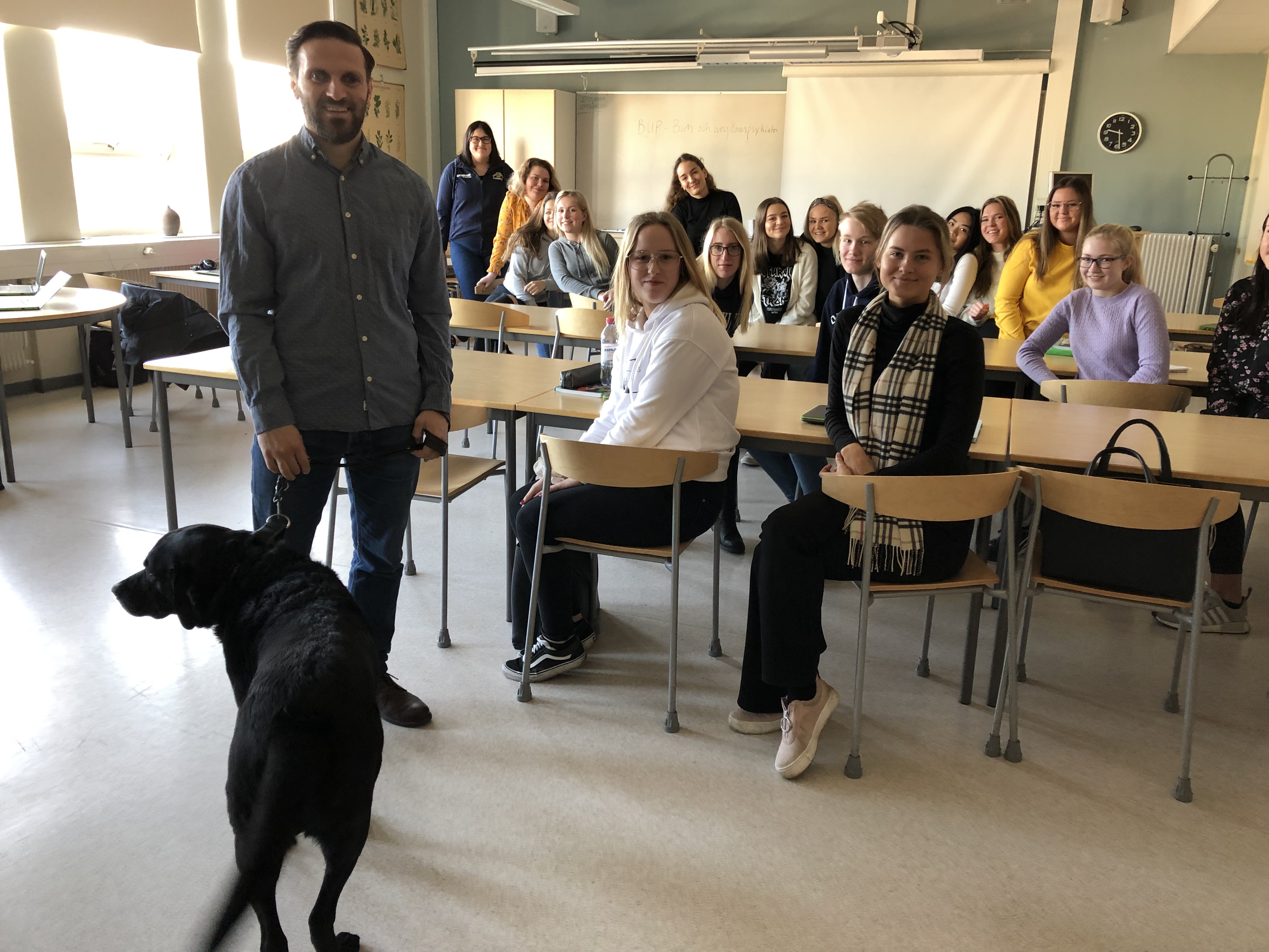 Fatmir och ledahunden Gibson står i ett klassrum tillsammans med ett 20-tal elever. 
