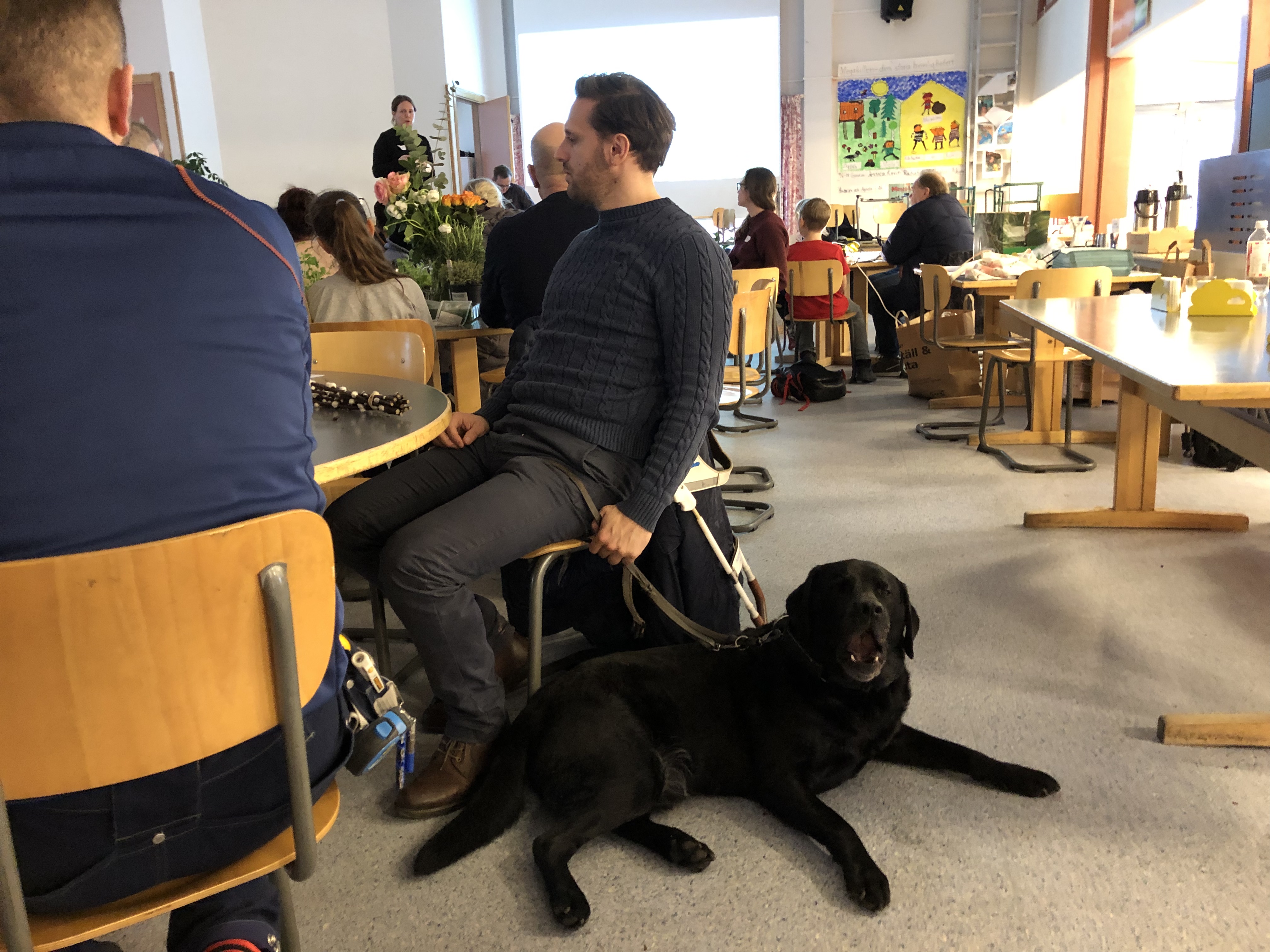 Fatmir och ledarhunden Gibson sitter vid ett bord och gibson ser trött ut och jässpar. Sinnernas trädgård, MKB Fastigheter. 