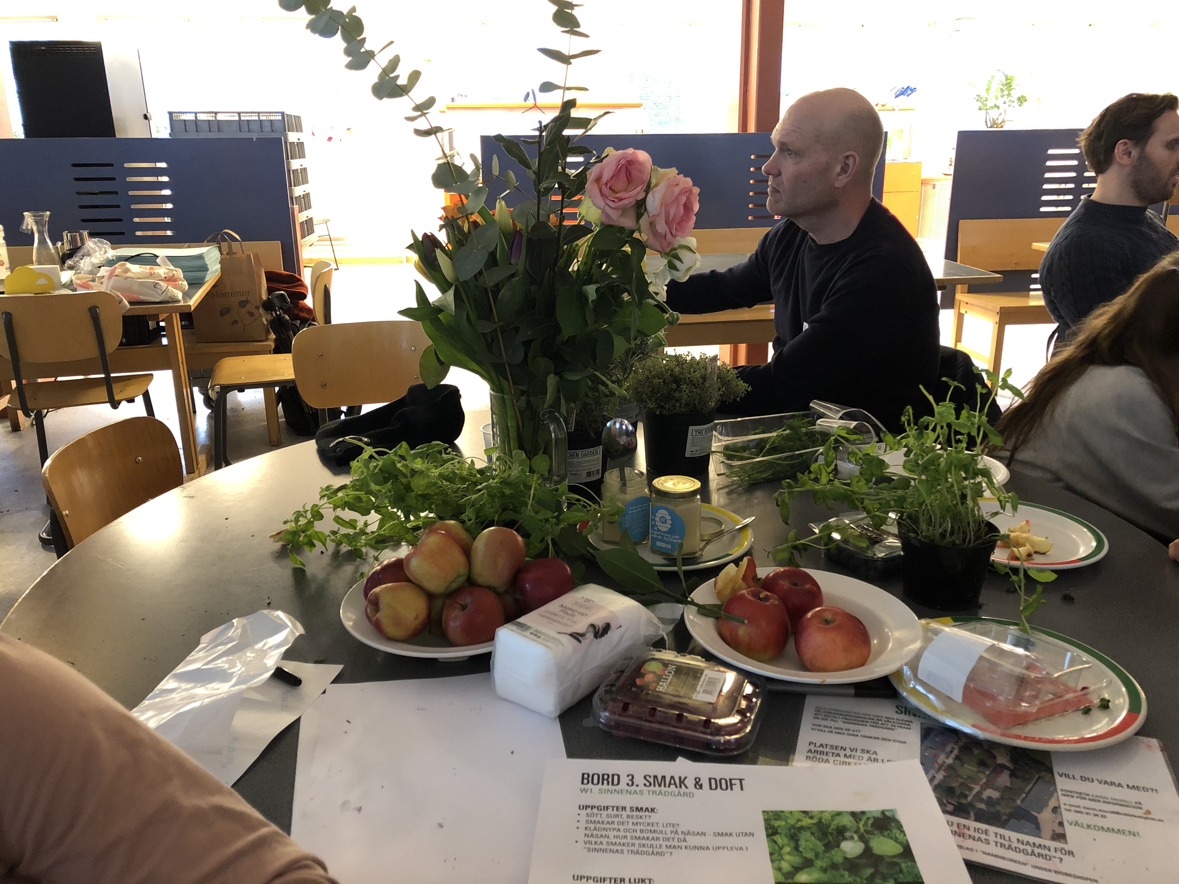 Fatmir och Gibson sitter med andra kring ett bord. På ett annat bord står det skålar med frukt och blommor. Sinnernas trädgård. MKB Fastigheter. 