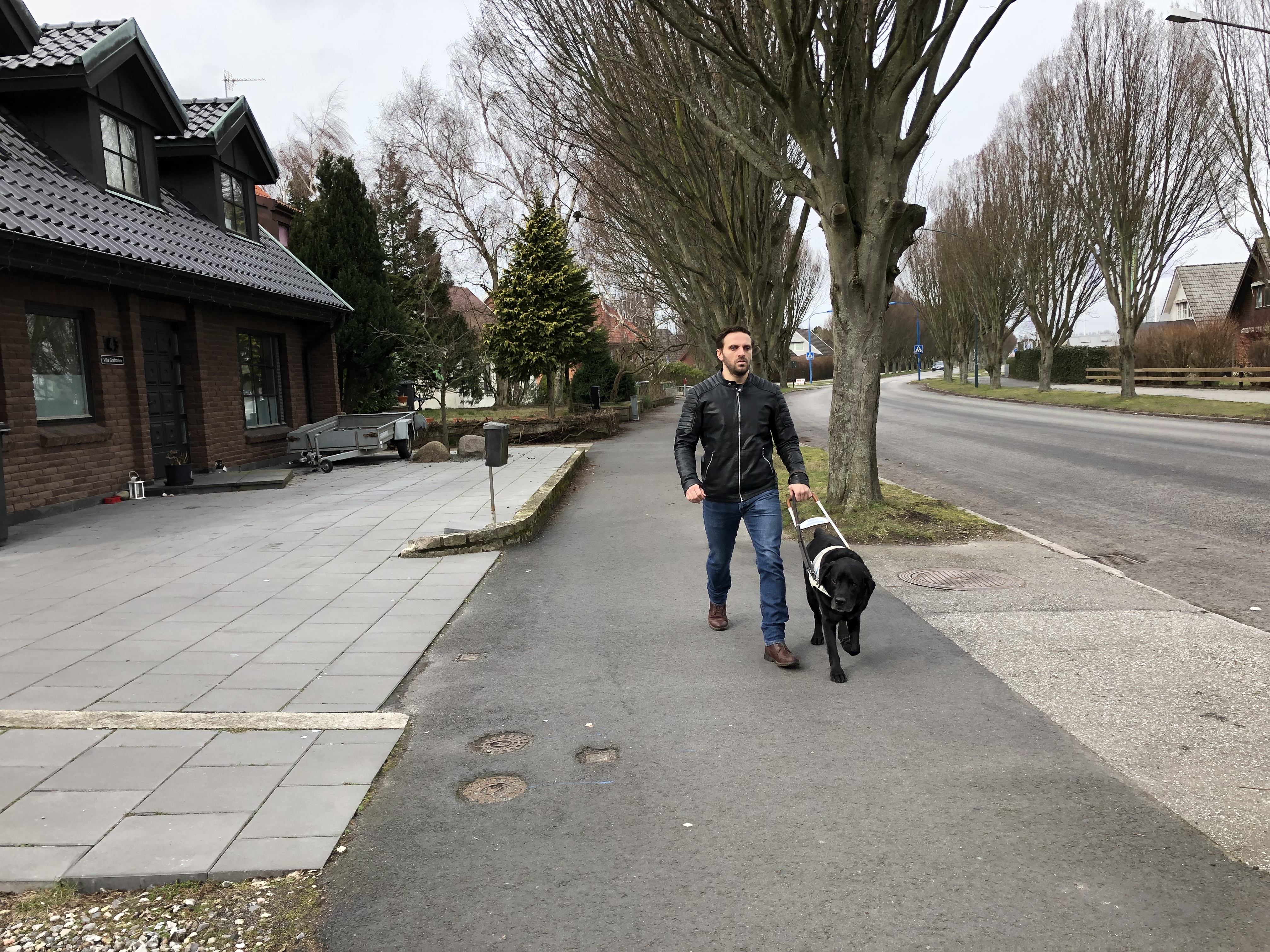 Fatmir är ute och går med gibson på trottoaren. Till vänster om honom finns en bilväg och till höger en gräsmatta, hus och träd. 