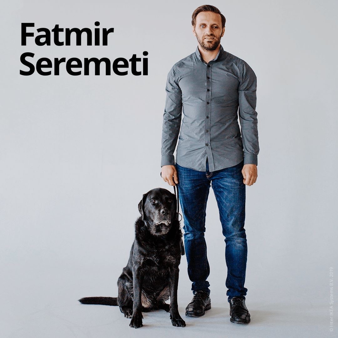 Fatmir Seremeti står på ett trägolv med sin ledarhund Gibson bredvid sig. Fatmir bär en mörkgrå skjorta och blå jeans och svarta finskor. 