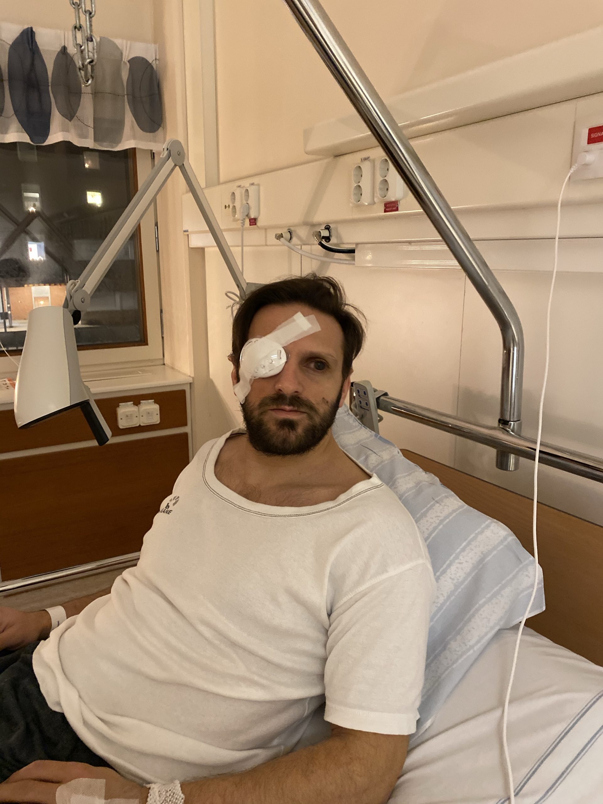 Fatmir Seremeti i en sjukhussäng i regionens sjukhuskläder på sig. Fatmir har ett bandage och plastkåpa för högerögat. 